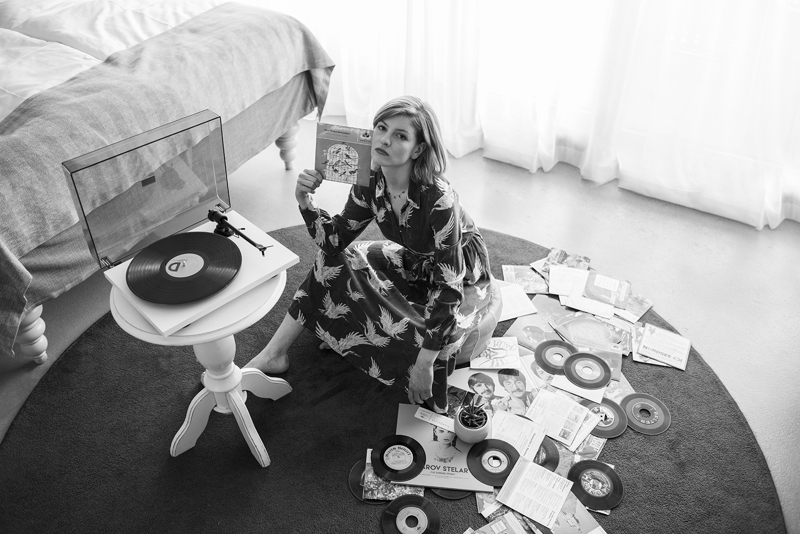 Tocadiscos T1 rodeado de discos en el suelo y una mujer sentada junto al tocadiscos sosteniendo un disco en el aire. Está ubicado en el dormitorio.